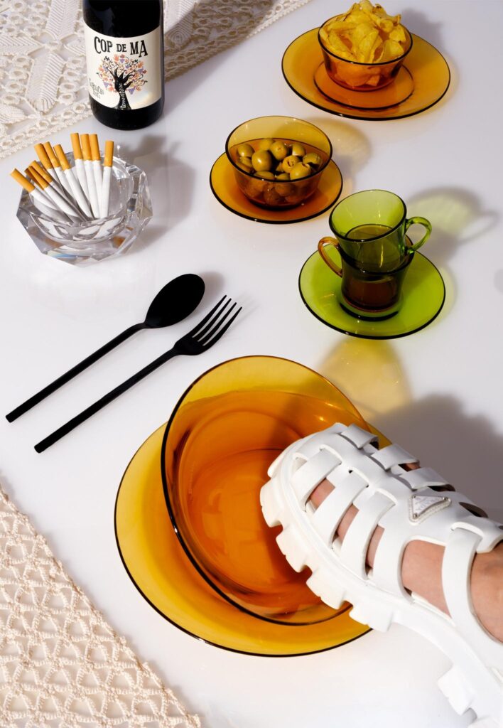 Composición de producto con vajilla Duralex ámbar, sandalia blanca Prada y luz dura sobre fondo blanco