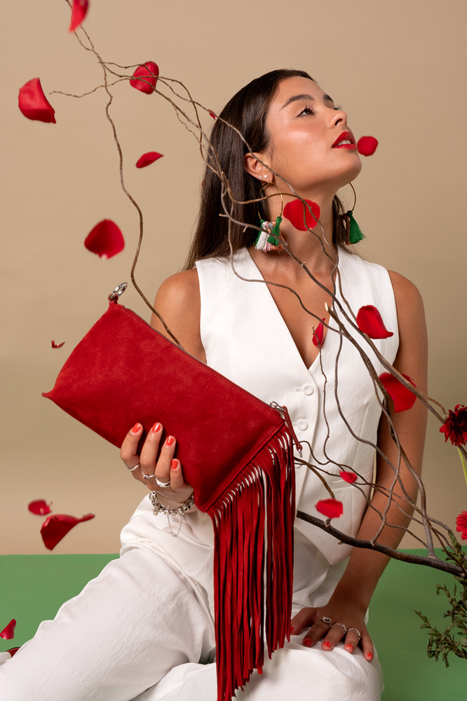 Mujer sosteniendo un bolso rojo con flecos, rodeada de pétalos de rosa y ramas decorativas.