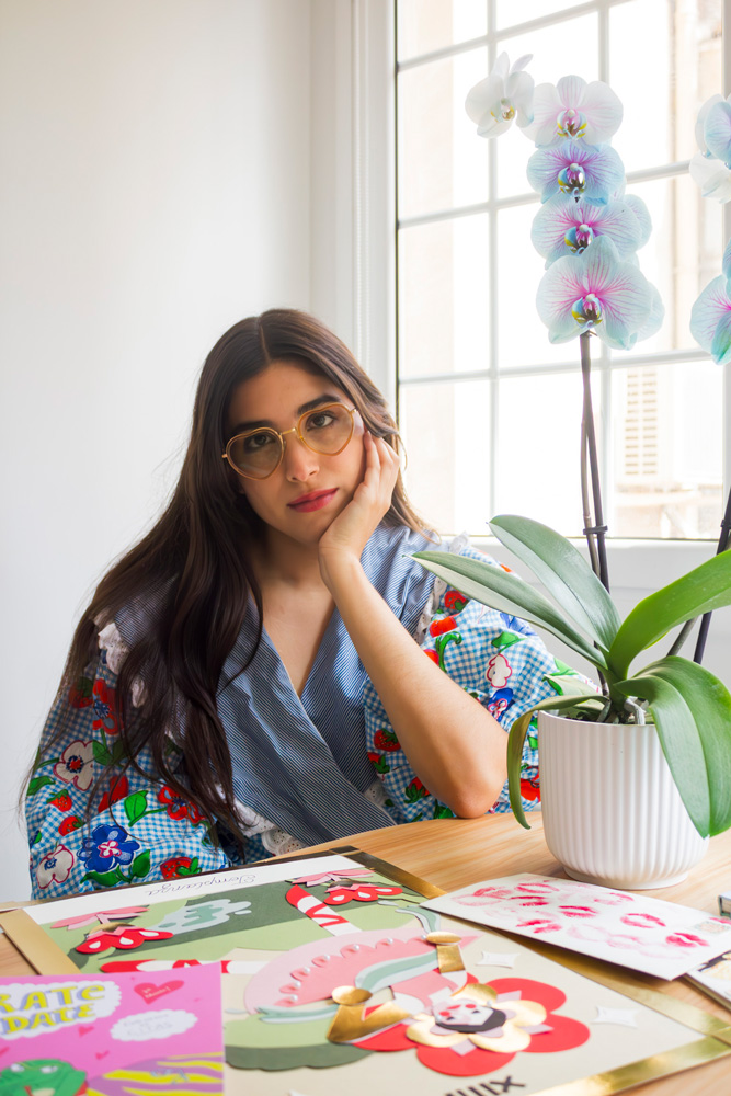 Mujer con gafas sentada frente a una mesa con ilustraciones coloridas y una orquídea a su lado.