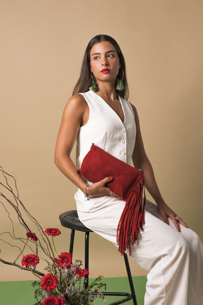 Mujer sosteniendo un bolso rojo con flecos, rodeada de pétalos de rosa y ramas decorativas.