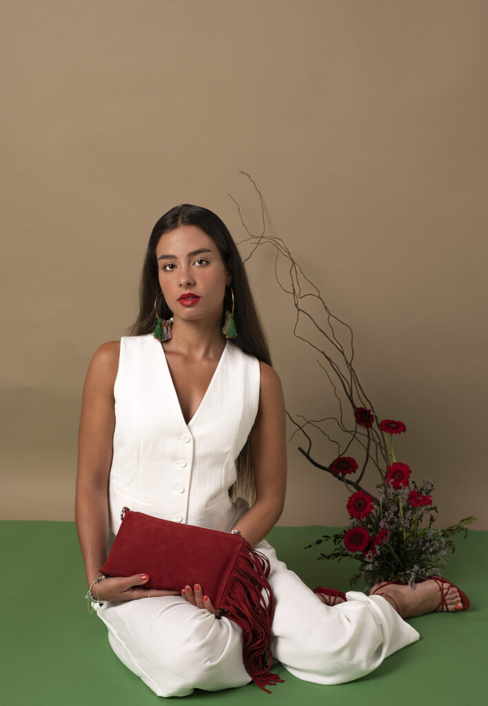 Mujer sosteniendo un bolso rojo con flecos, rodeada de pétalos de rosa y ramas decorativas.