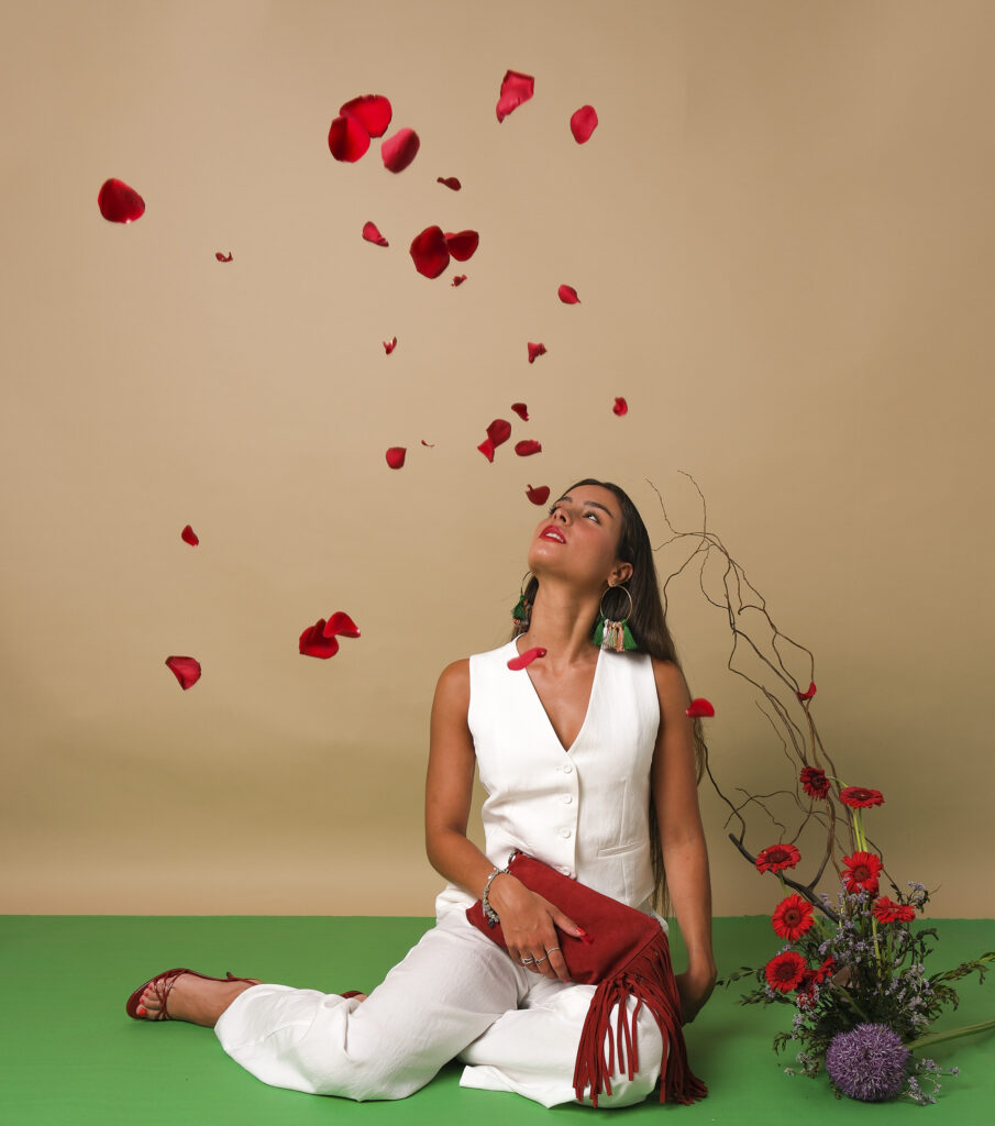 Mujer sosteniendo un bolso rojo con flecos, rodeada de pétalos de rosa y ramas decorativas.
