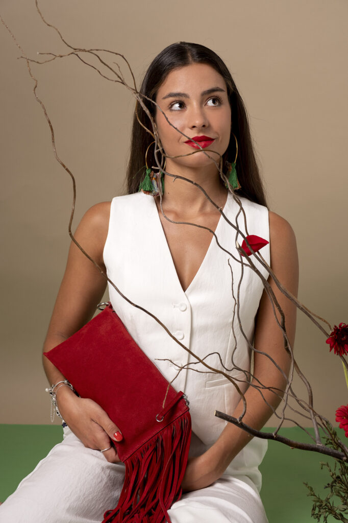 Mujer sosteniendo un bolso rojo con flecos, rodeada de pétalos de rosa y ramas decorativas.