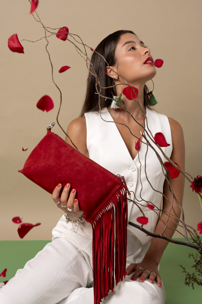 Mujer sosteniendo un bolso rojo con flecos, rodeada de pétalos de rosa y ramas decorativas.
