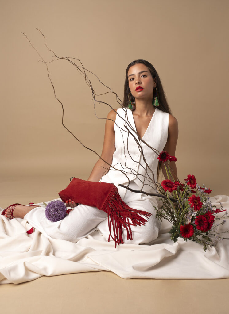 Mujer sosteniendo un bolso rojo con flecos, rodeada de pétalos de rosa y ramas decorativas.